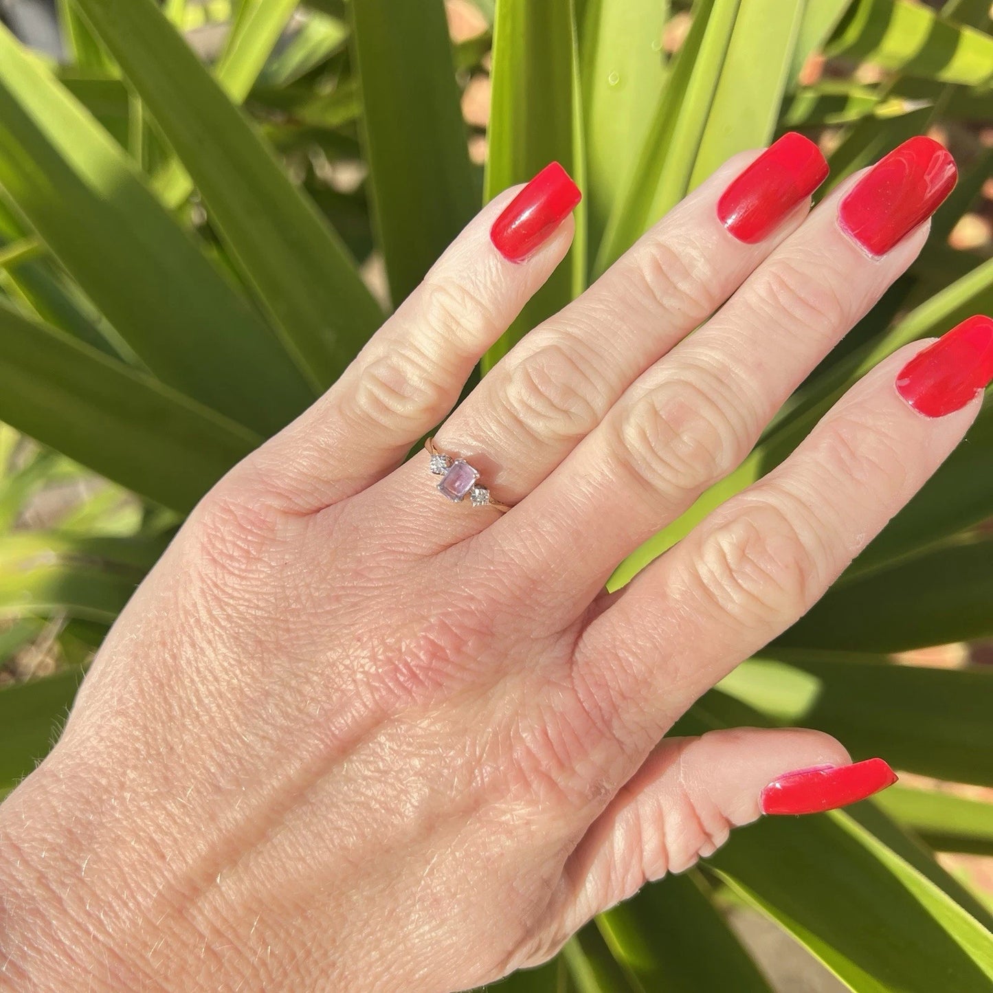 Hand with red nail polish wearing a purple amethyst gemstone and diamonds ring against a green leafy background