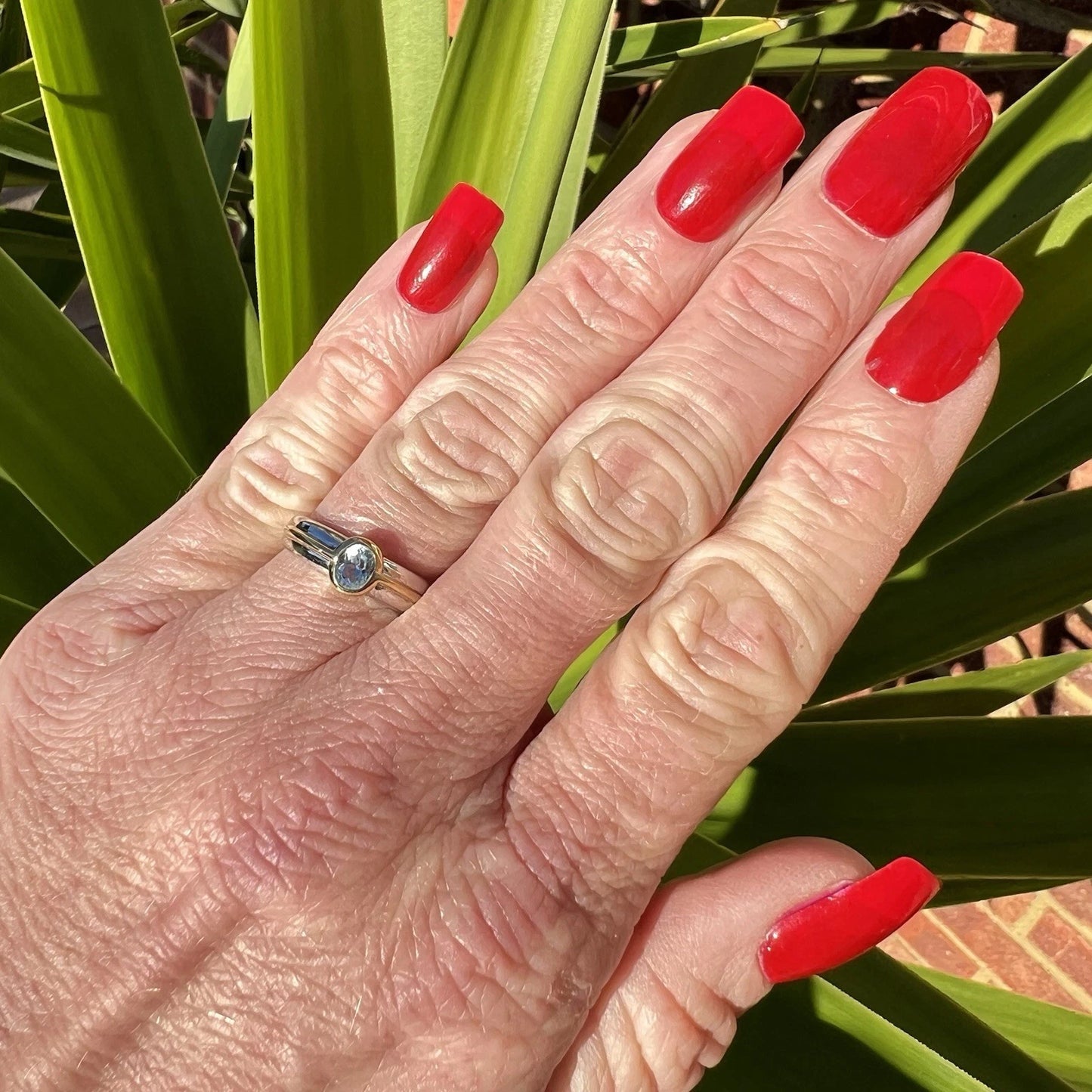Hand with red nail polish wearing a gold ring with a blue topaz against a green leafy background