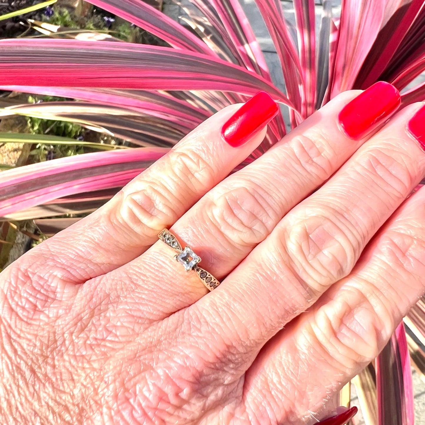 Hand with red nail polish wearing ring with pink and green plant in background