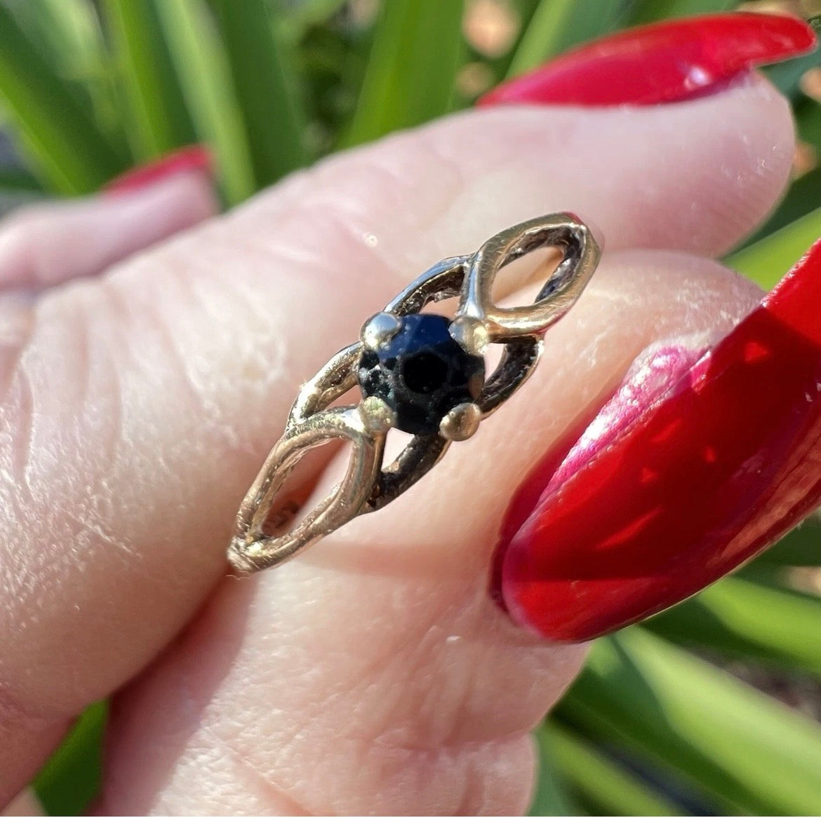 Gold ring with a blue gemstone on a hand with red nail polish against a blurred green background