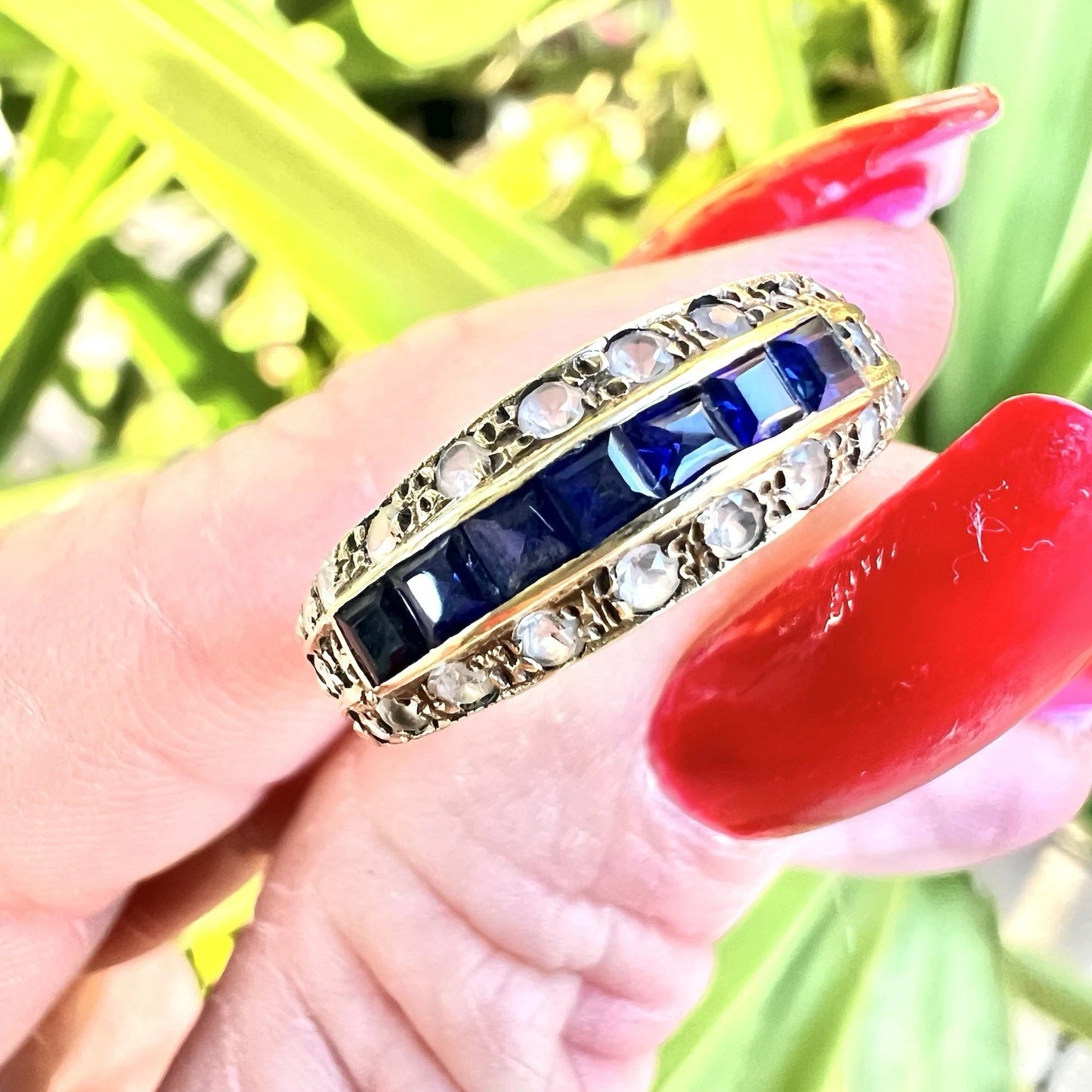 Gold ring with blue sapphire gemstones being held by a hand with red nail polish against a blurred green background