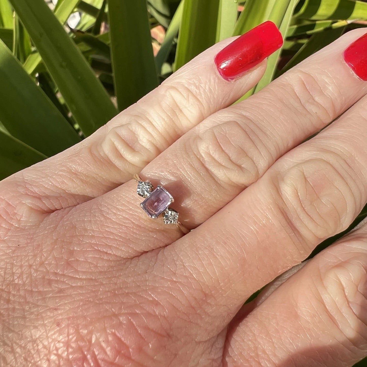 Hand wearing a ring with a purple amethyst gemstone and diamonds against a green leafy background