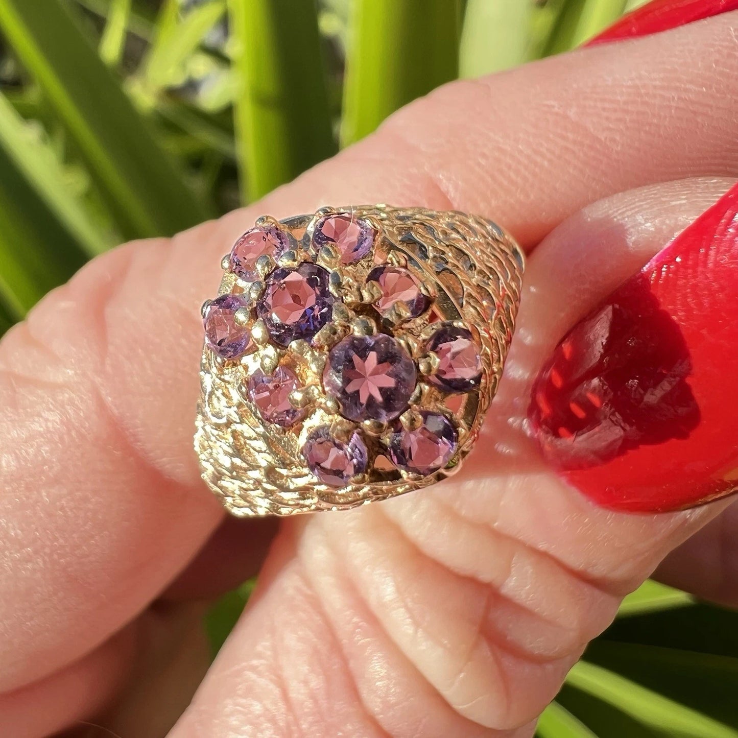 Gold ring with purple amethyst gemstones held by a hand with red nail polish against a green background.