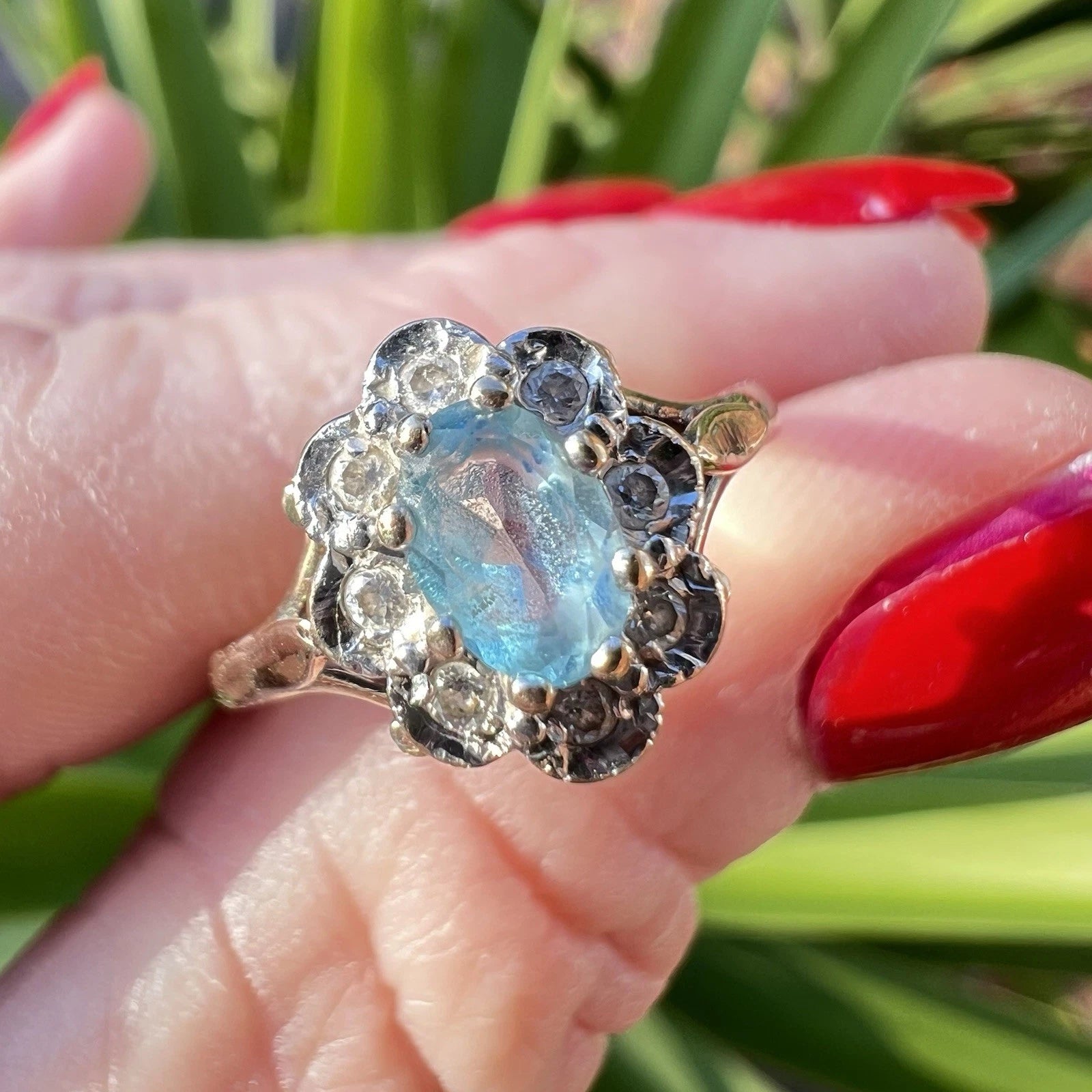 Gold ring with a blue topaz gemstone and diamond halo held by a hand with red nail polish against a green leafy background.