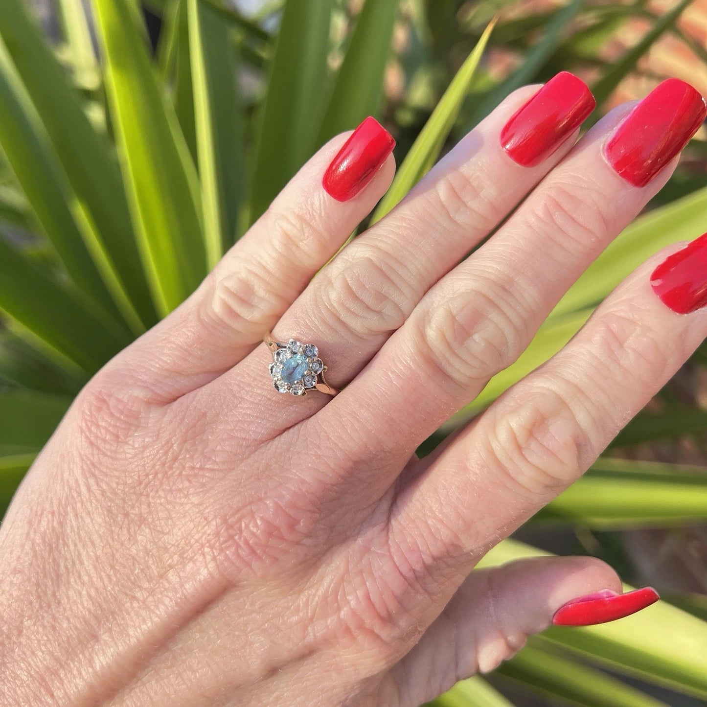 Hand with red nail polish wearing a ring with a blue topaz gemstone and diamond halo against a green leafy background