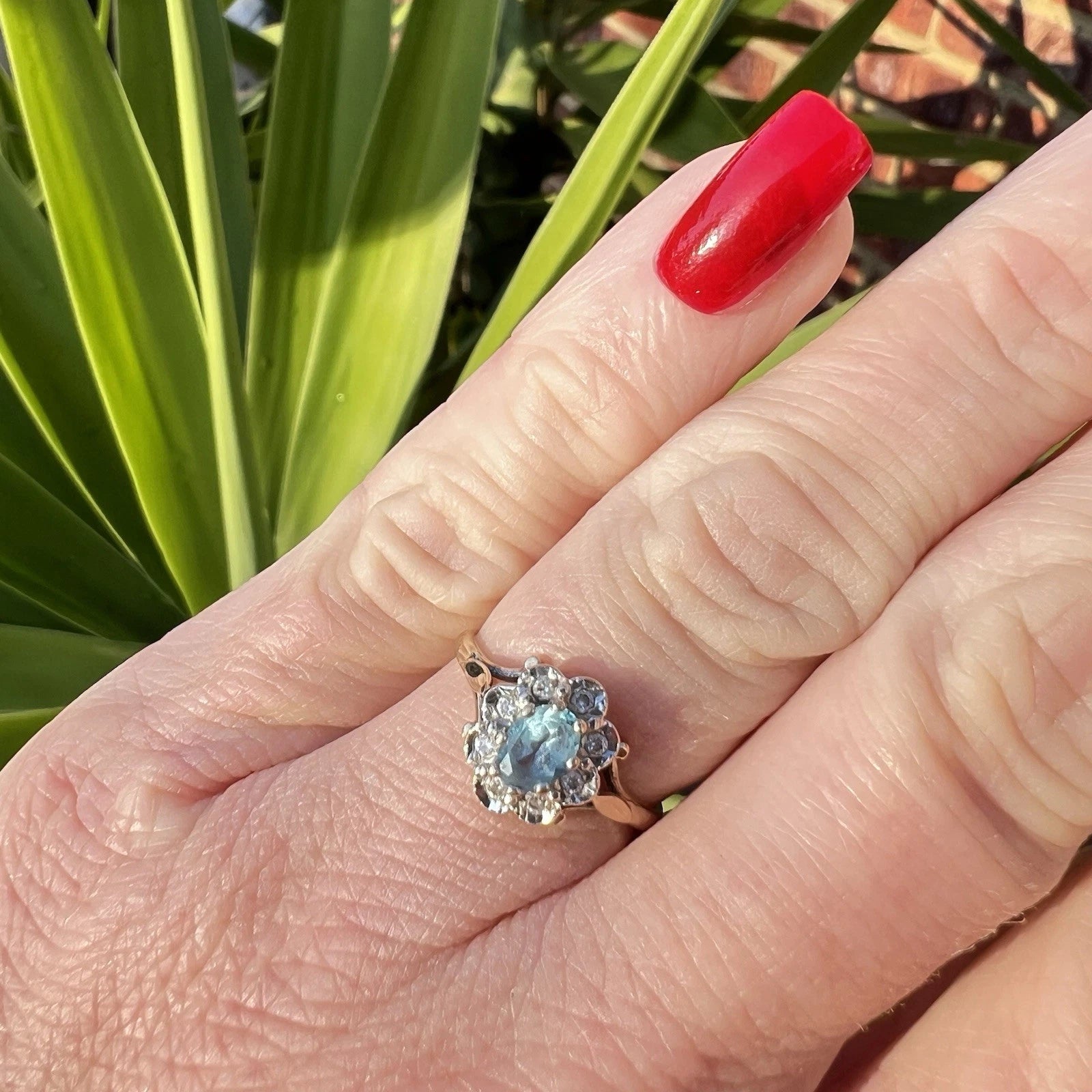 Hand wearing a decorative ring with a blue topaz gemstone and diamond halo against a green plant background