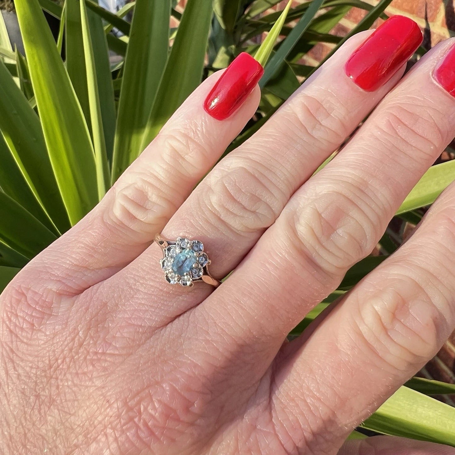 Hand wearing a ring with a blue topaz and diamond halo against a green leafy background