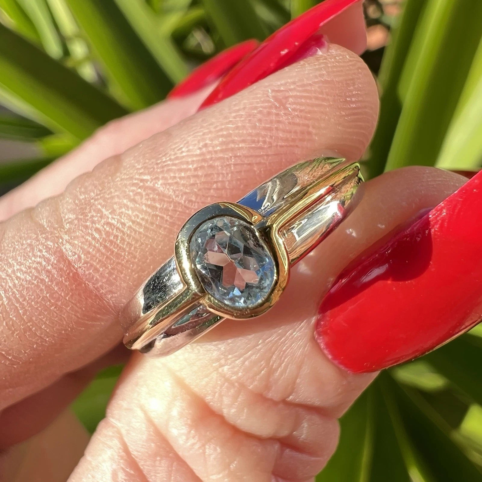 Gold ring with a blue topaz gemstone on a finger with red nail polish against a green background