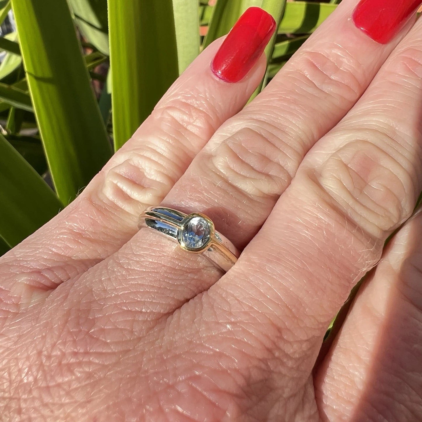 Close-up of a hand wearing a gold ring with a blue topaz gemstone, against a green leafy background.