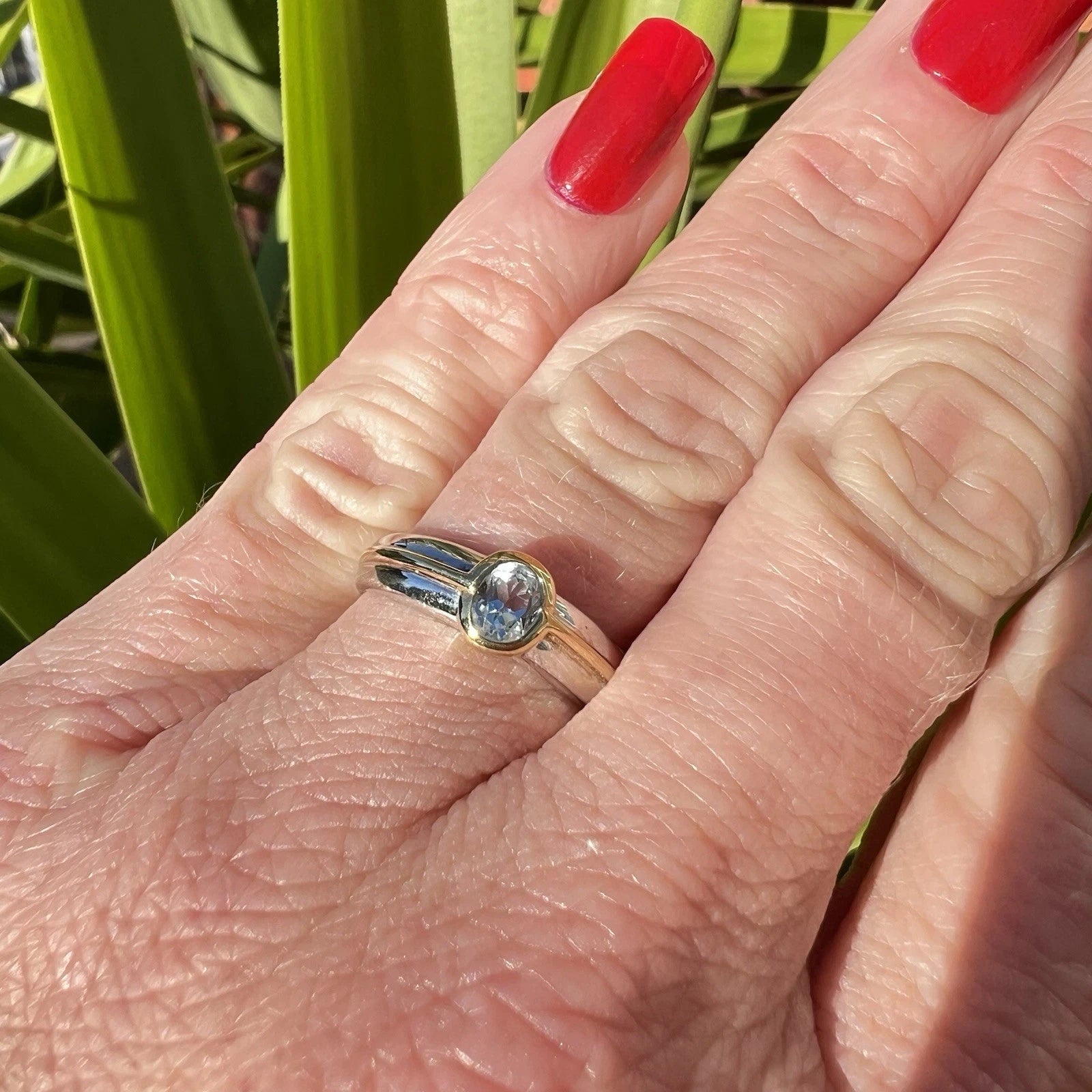 Close-up of a hand wearing a gold ring with a blue topaz gemstone, against a green leafy background.