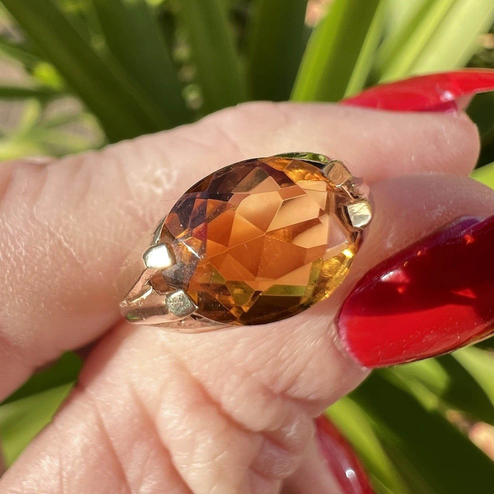 Ring with a large orange citrine gemstone held by a hand with red nail polish against a green background