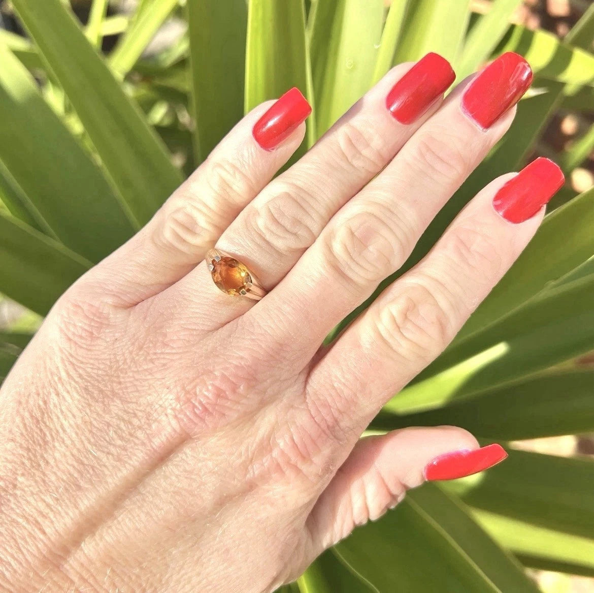 Hand with red nail polish wearing an citrine ring against a green leafy background