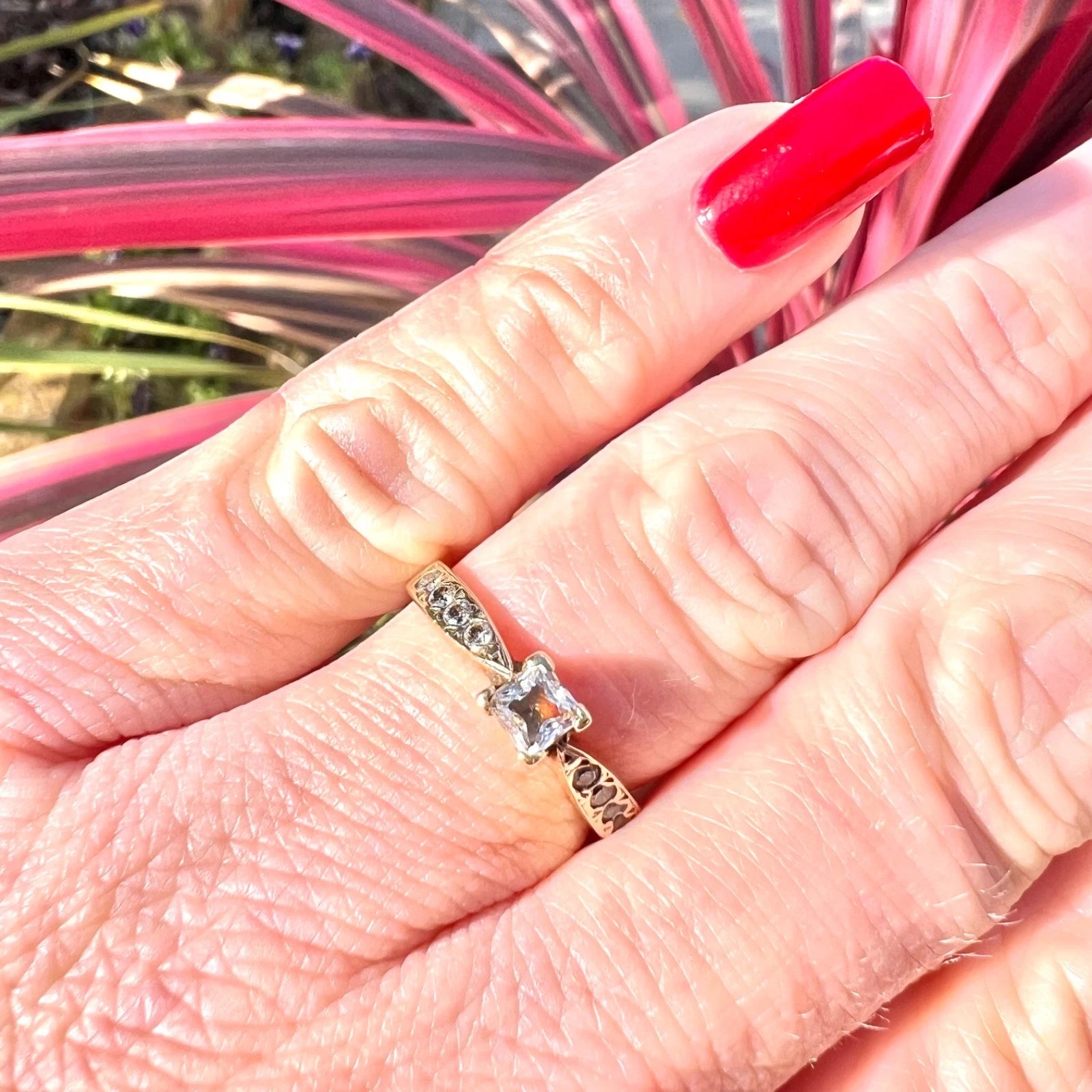 Close-up of a hand wearing a gold solitaire ring with a cubic zirconia , set against a pink and green plant background.