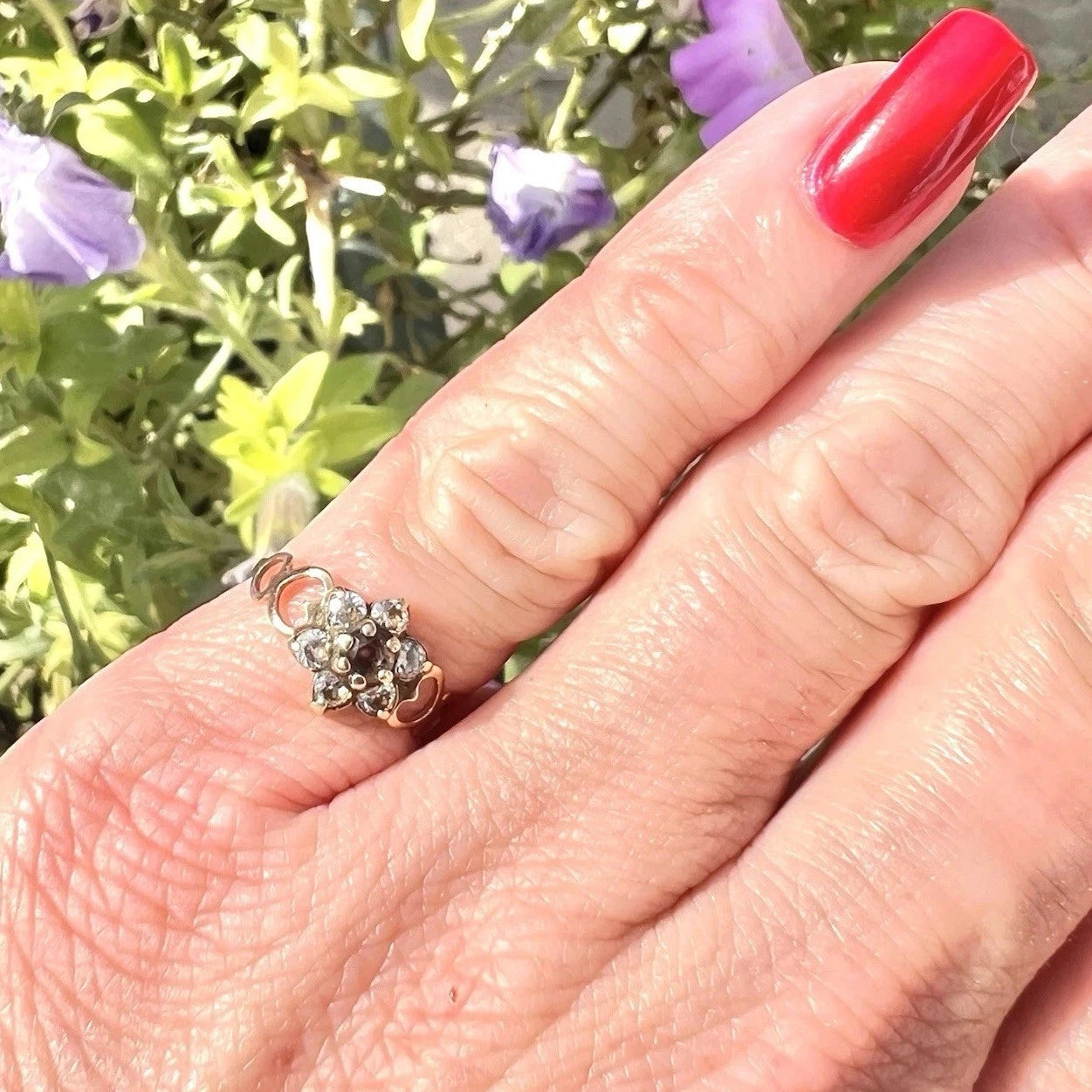 Hand wearing a decorative ring with red nail polish against a blurred floral background