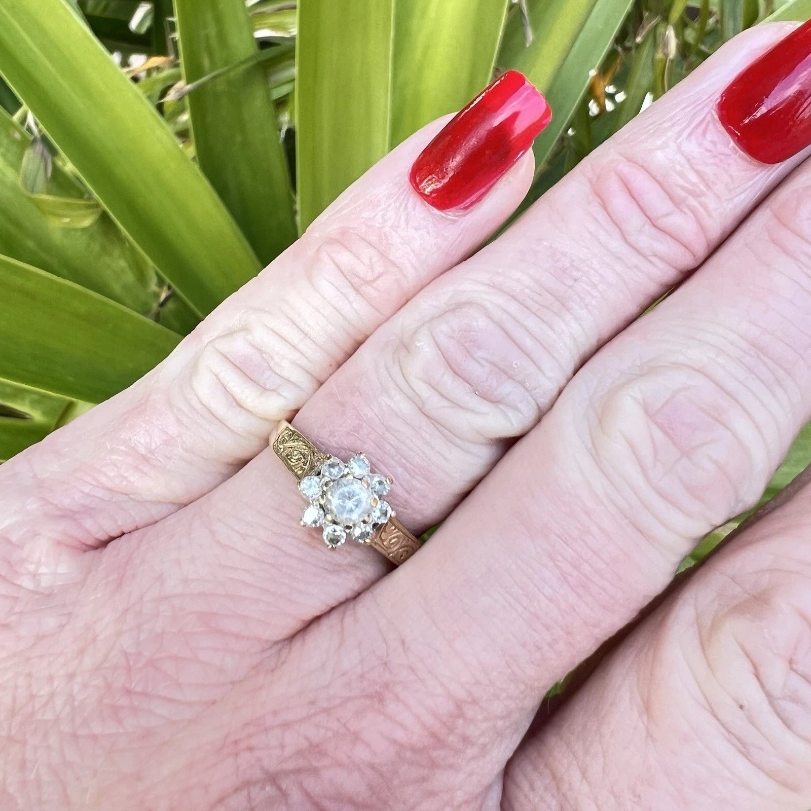 Hand wearing a gold ring with a cubic zirconia flower cluster, red nail polish against a green leafy background
