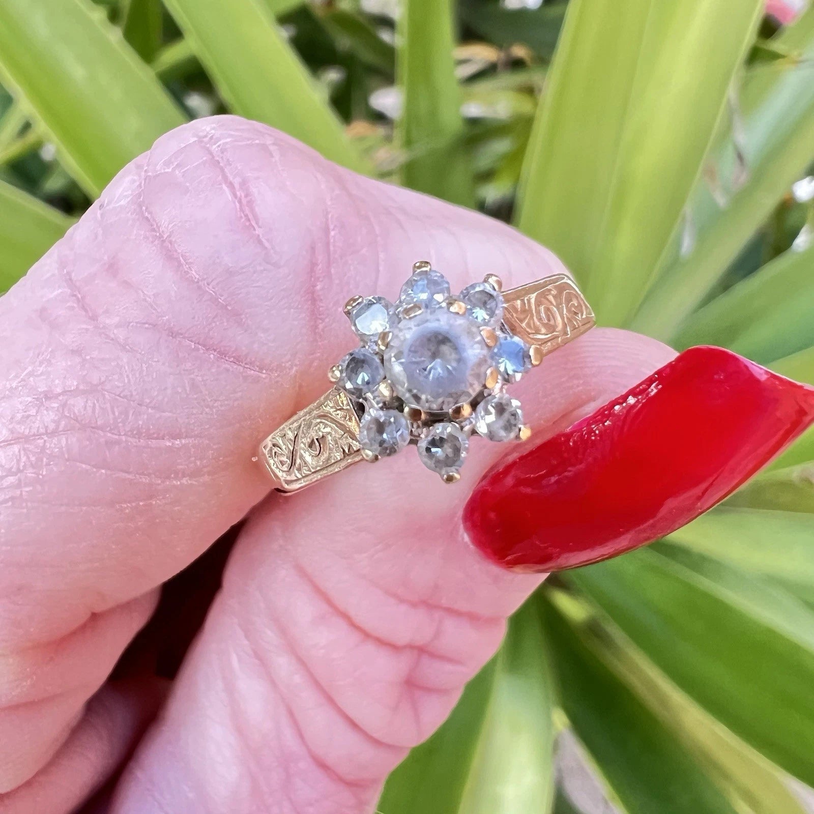 Gold ring with a central gemstone held by a hand with red nail polish against a green leafy background