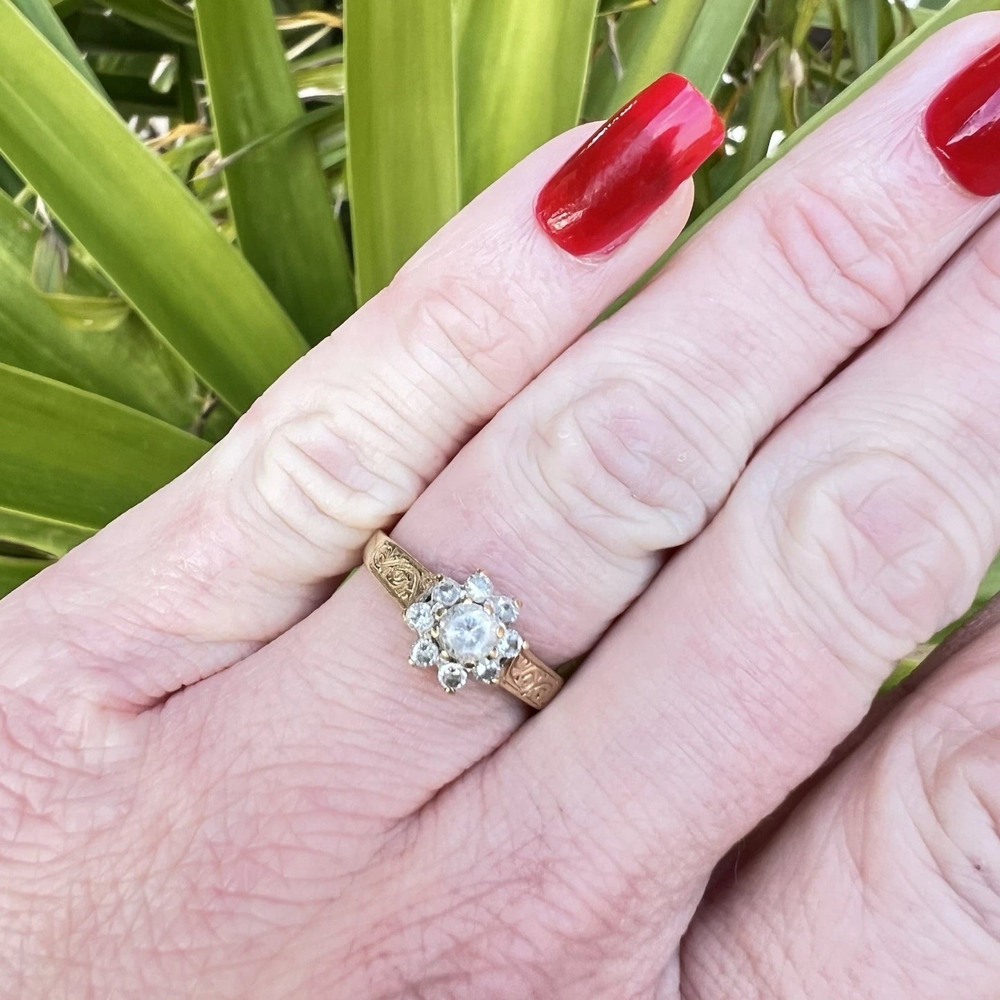 Hand wearing a cubic zirconia ring with red nail polish against a green leafy background