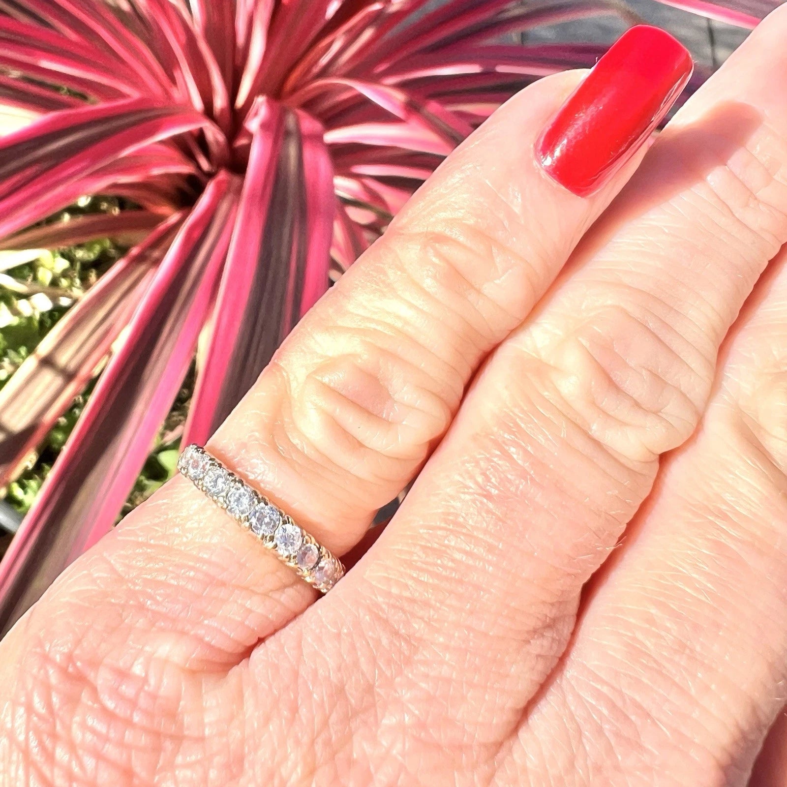 Close-up of a hand with a cubic zirconia ring and red nail polish against a pink plant background