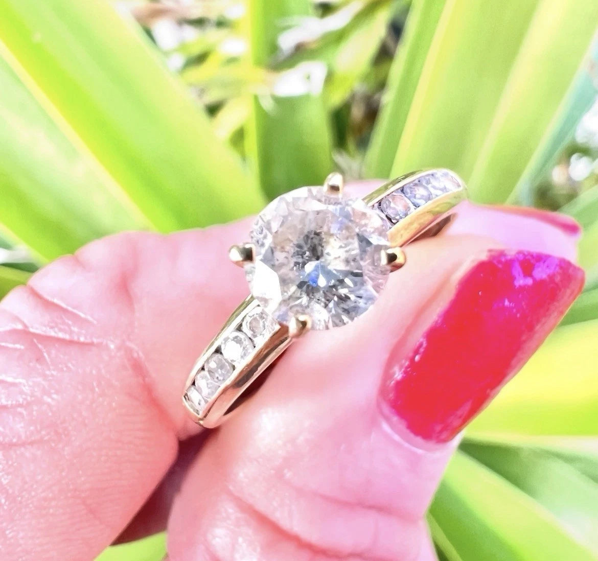 Gold ring with a large CZ held by a hand with red nail polish against a green plant background