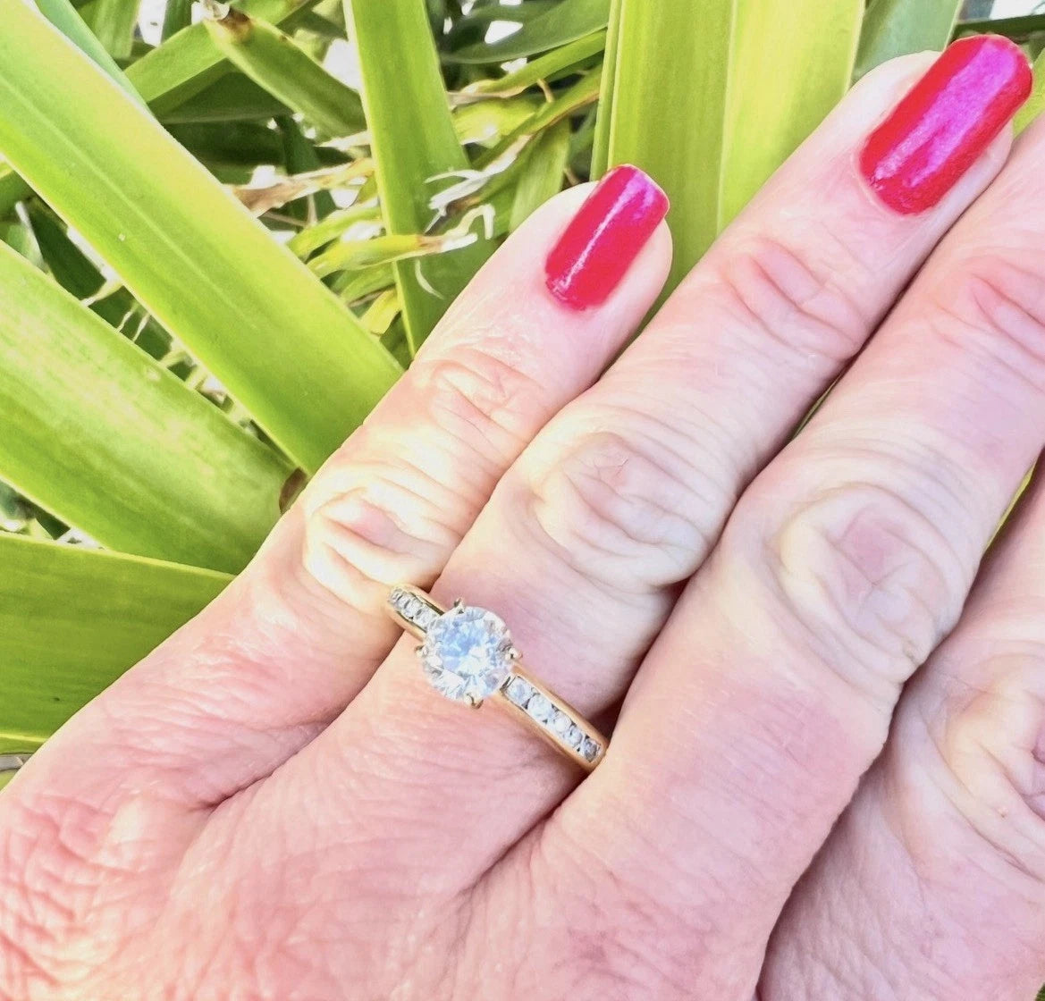 Hand wearing a gold CZ ring with red nail polish against a green leafy background