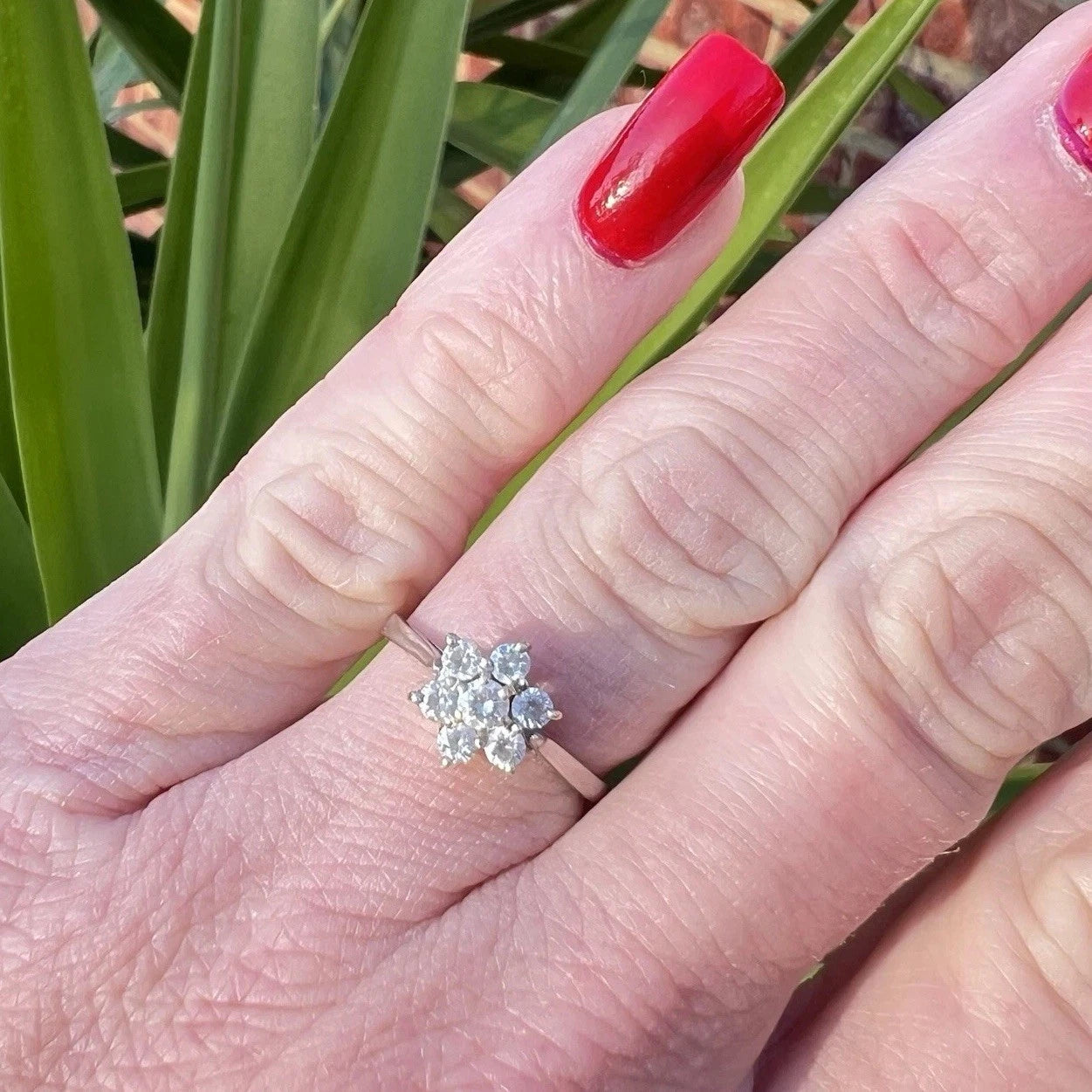 Hand wearing a white gold ring with cubic zirconia cluster flower setting against a green leafy background