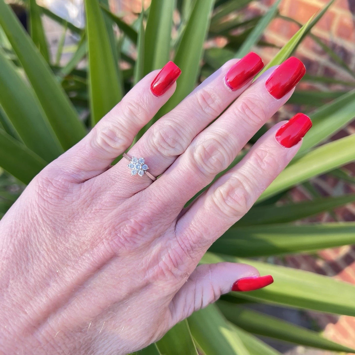 Hand with red nails wearing a cubic zirconia ring against a green leafy background