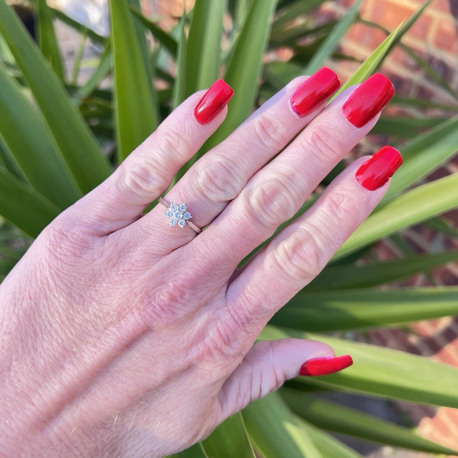 Hand with red nails wearing a cubic zirconia ring against a green leafy background