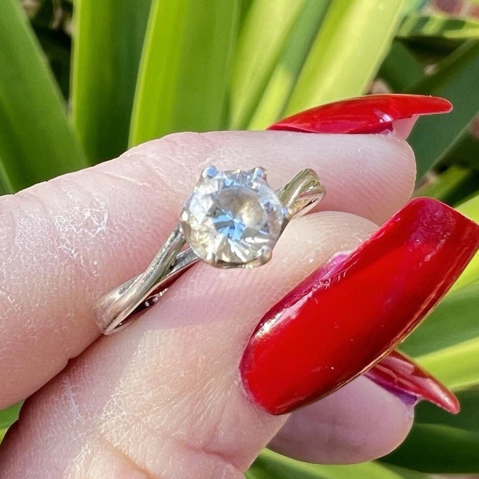 White Gold ring with a large cubic zirconia on a finger with red nail polish against a green leafy background