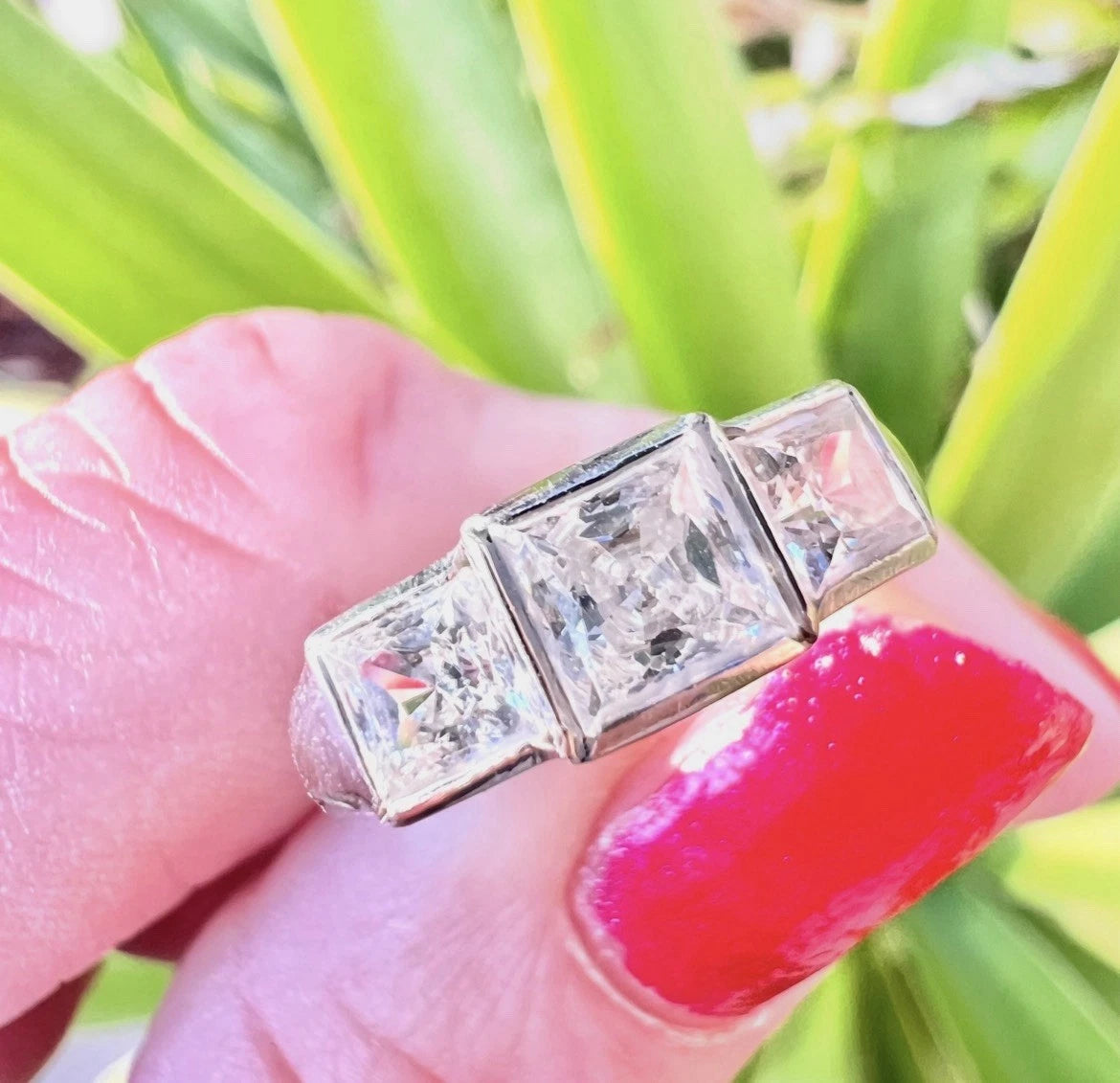 White fold ring with three clear gemstones held between fingers against a green leafy background