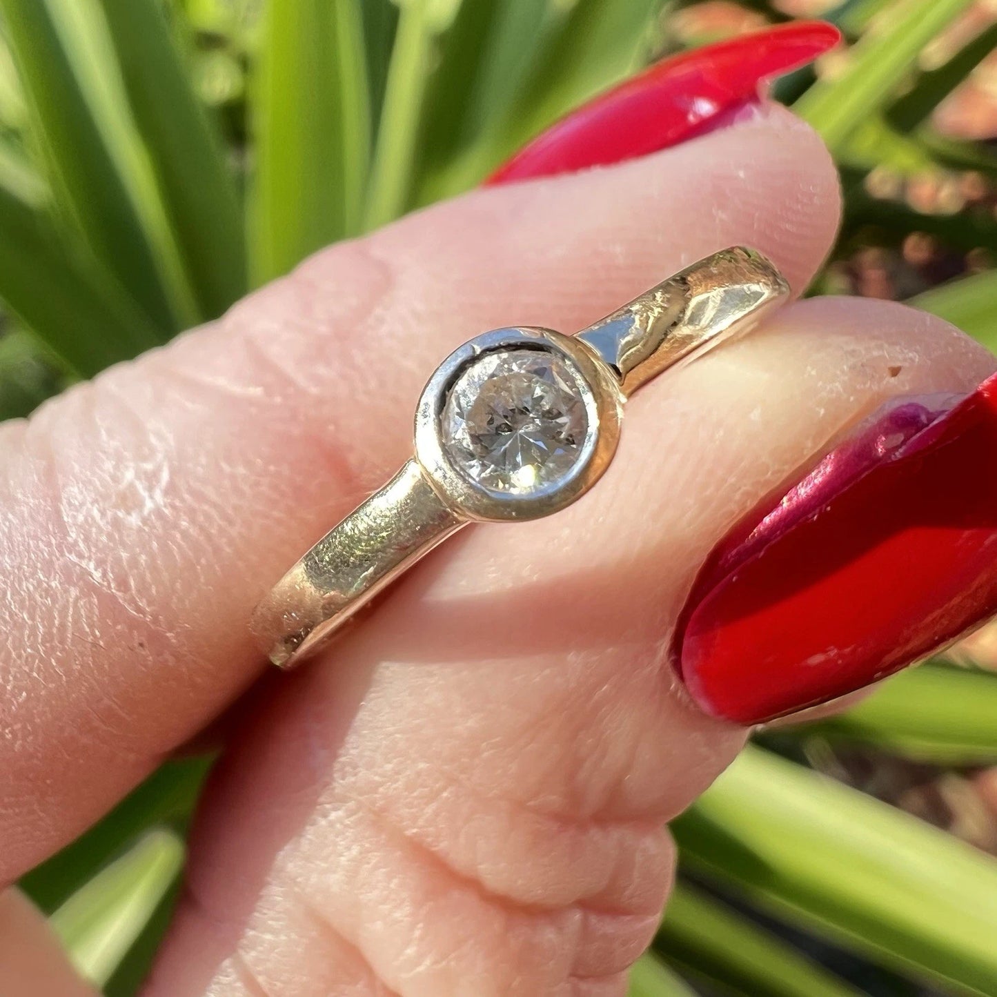 Gold ring with a cubic zirconia being held by fingers with red nail polish against a green background