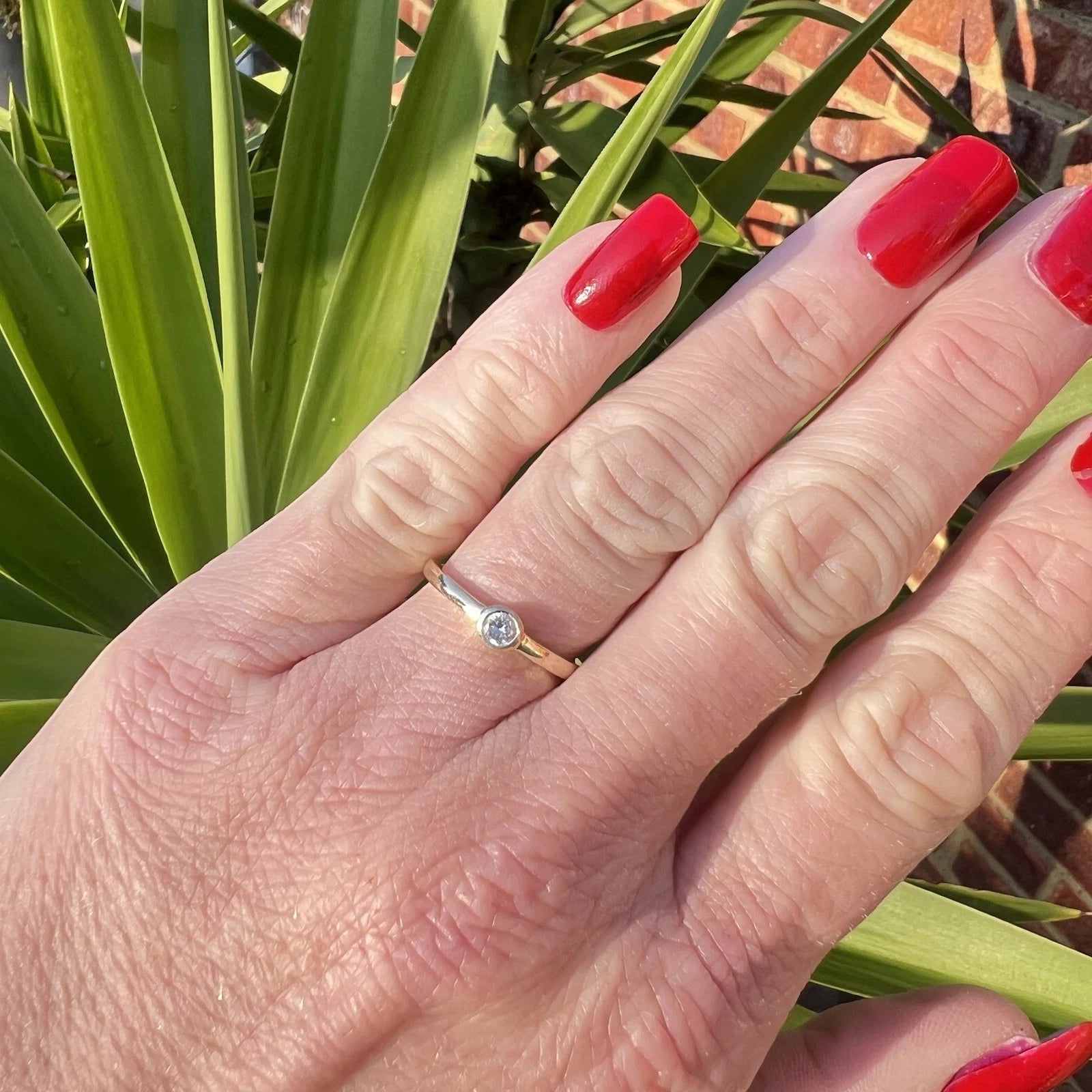 Hand with red nail polish wearing a gold cubic zirconia solitaire ring against a green leafy background
