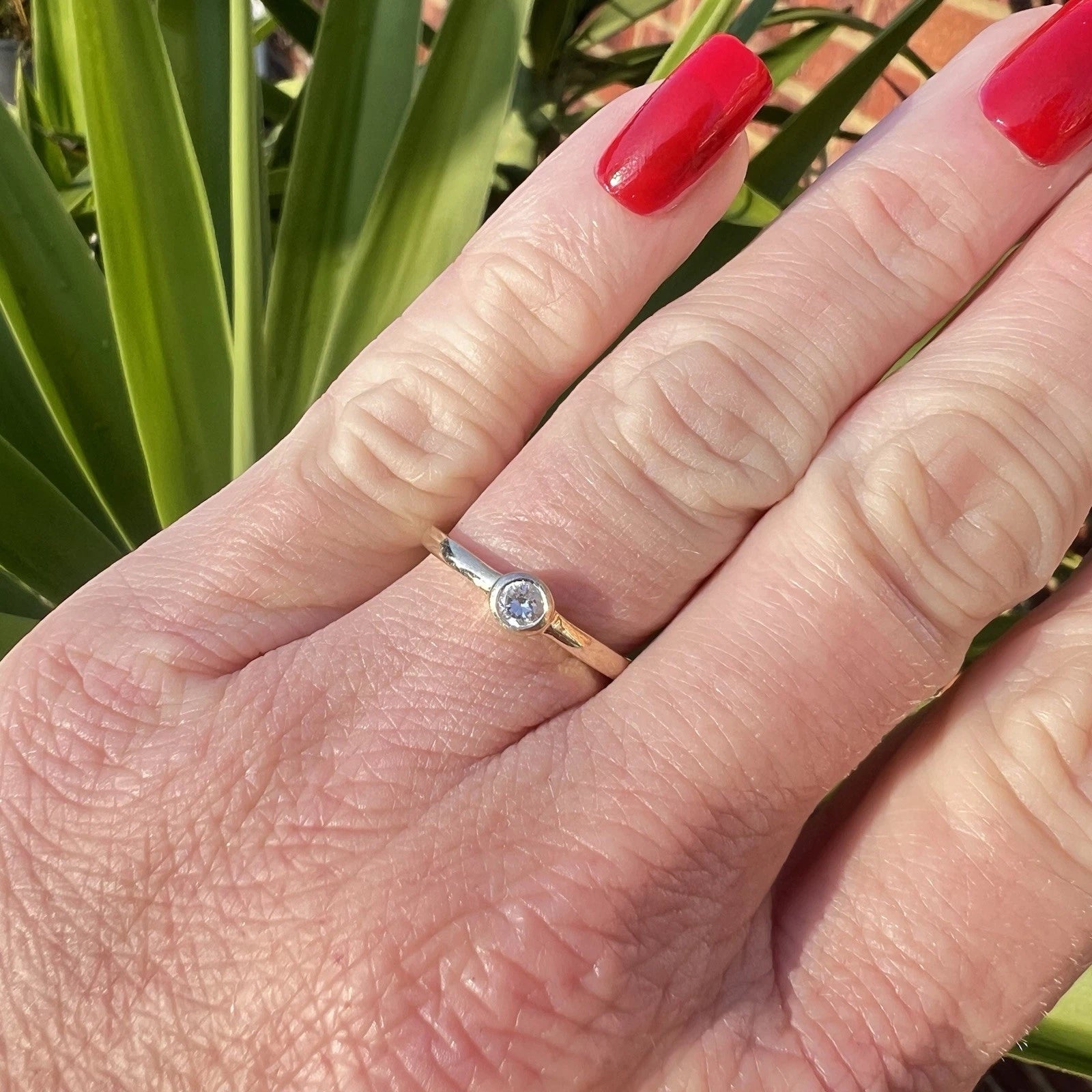 Hand wearing a gold ring with a cubic zirconia on a blurred green plant background