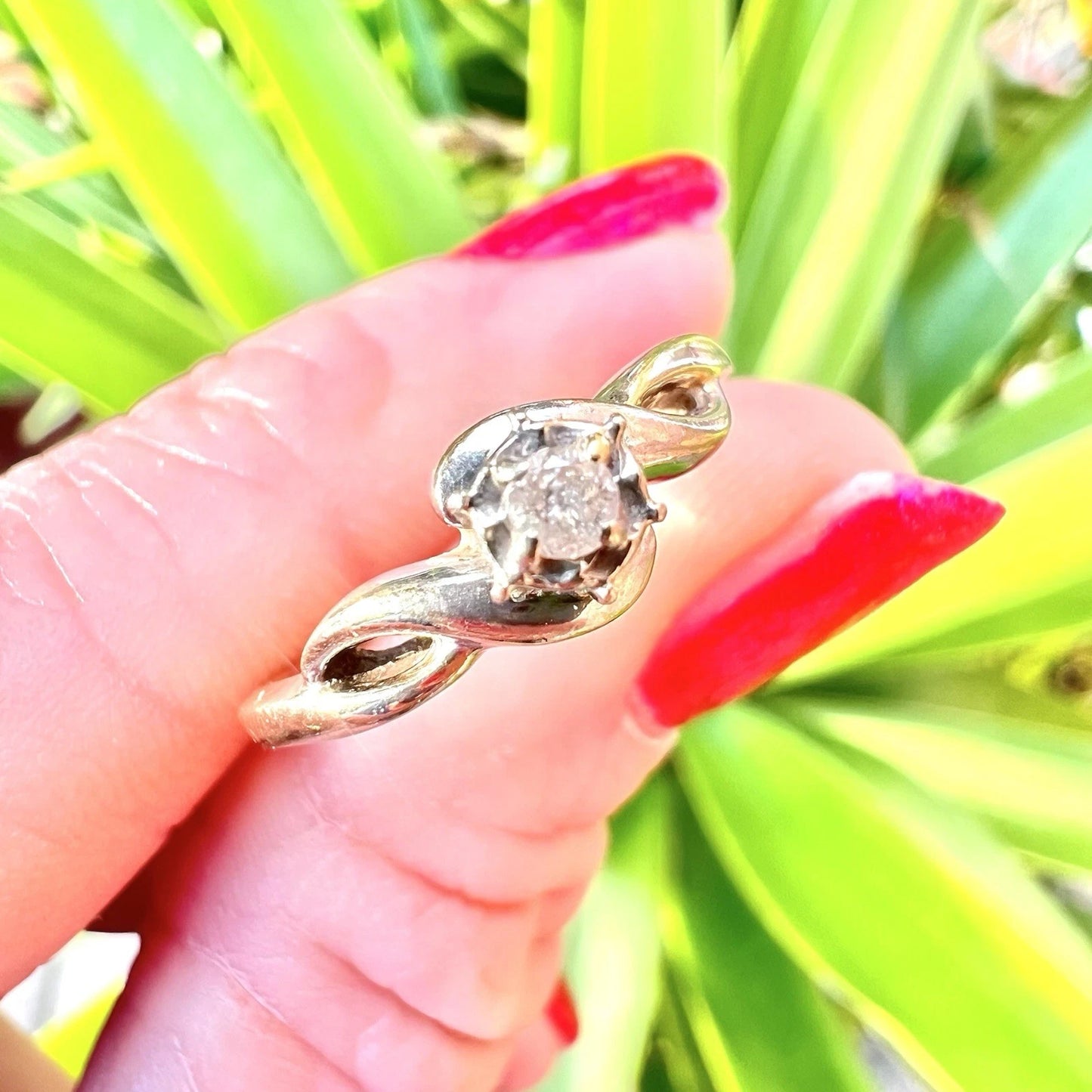Gold ring with a diamond held by a hand with red nail polish against a green leafy background