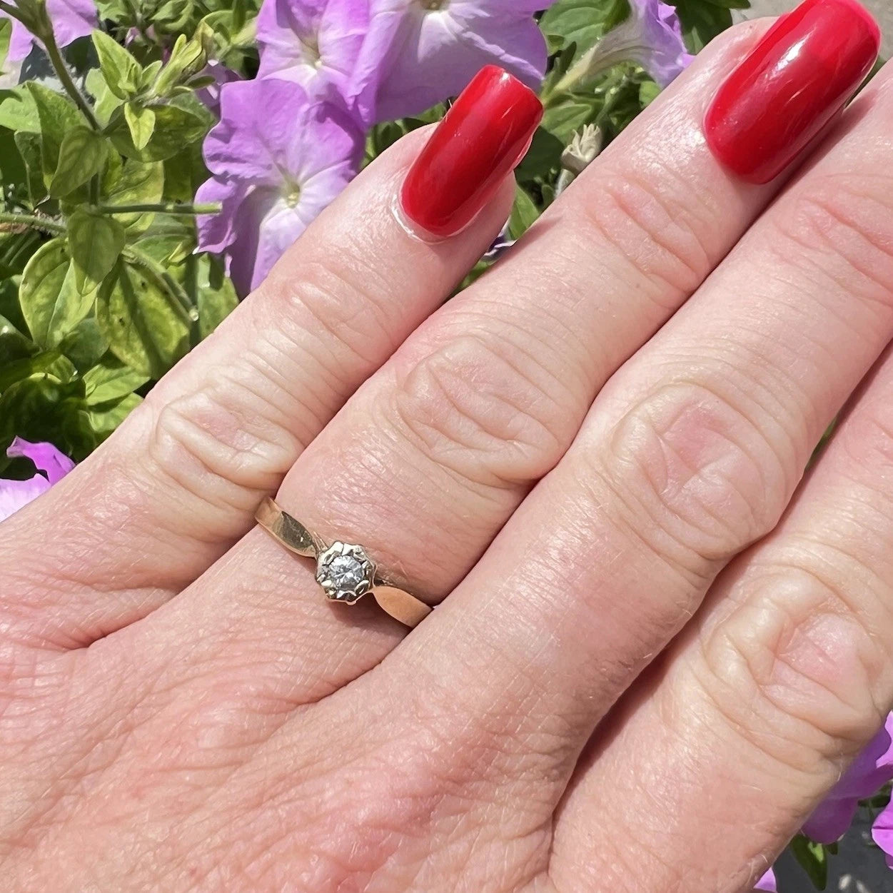 Hand with red nail polish wearing a fold diamond solitaire ring against a floral background