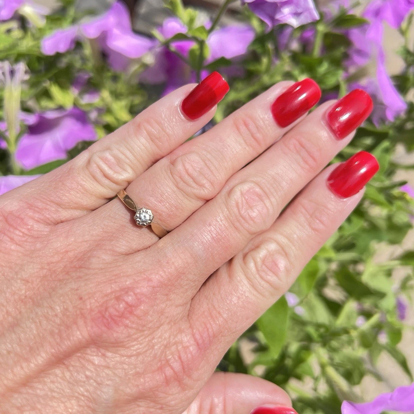 Hand with red nail polish wearing a gold ring with a diamond against a blurred floral background