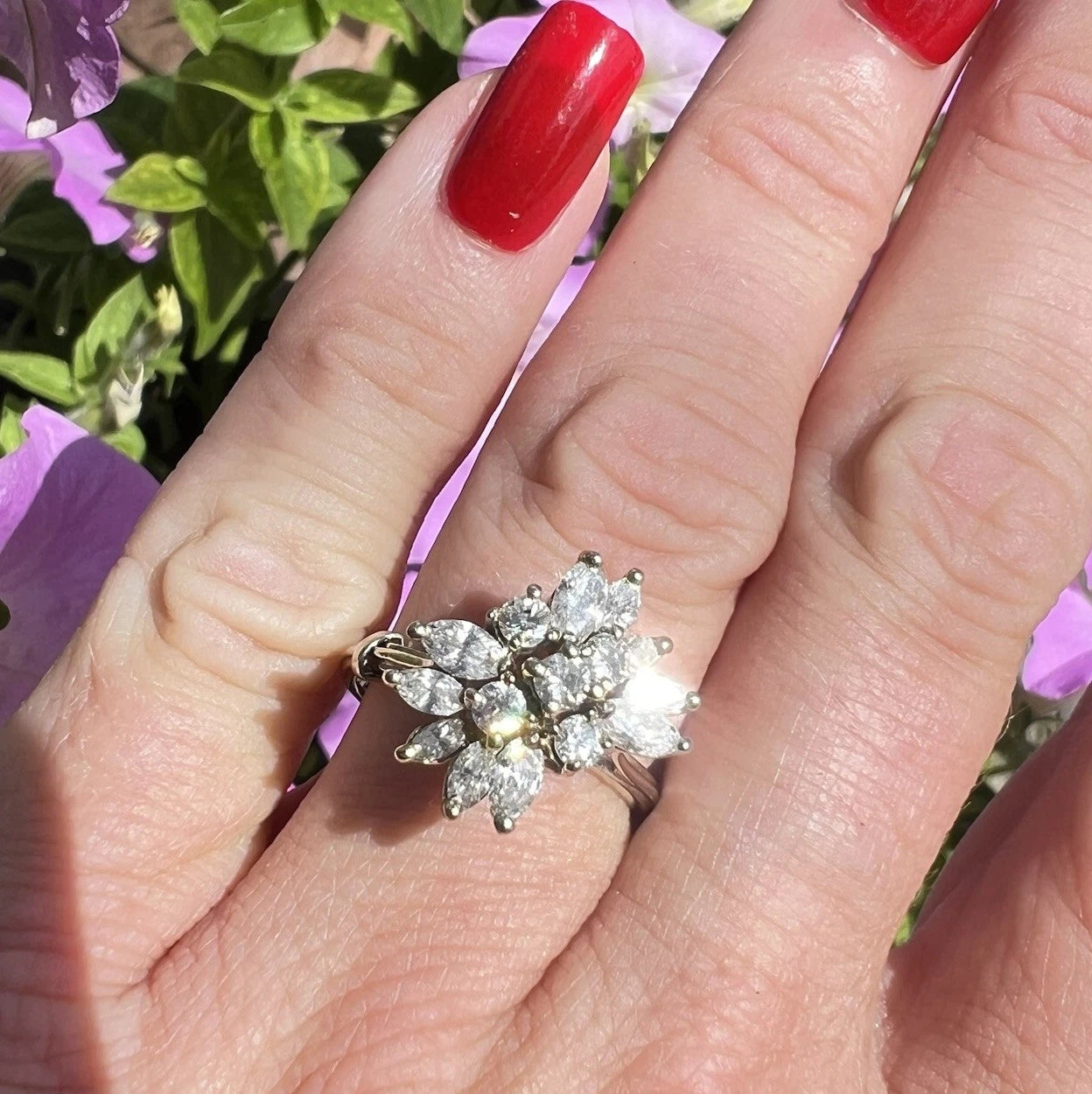 Hand wearing a diamond ring with red nail polish against a blurred floral background