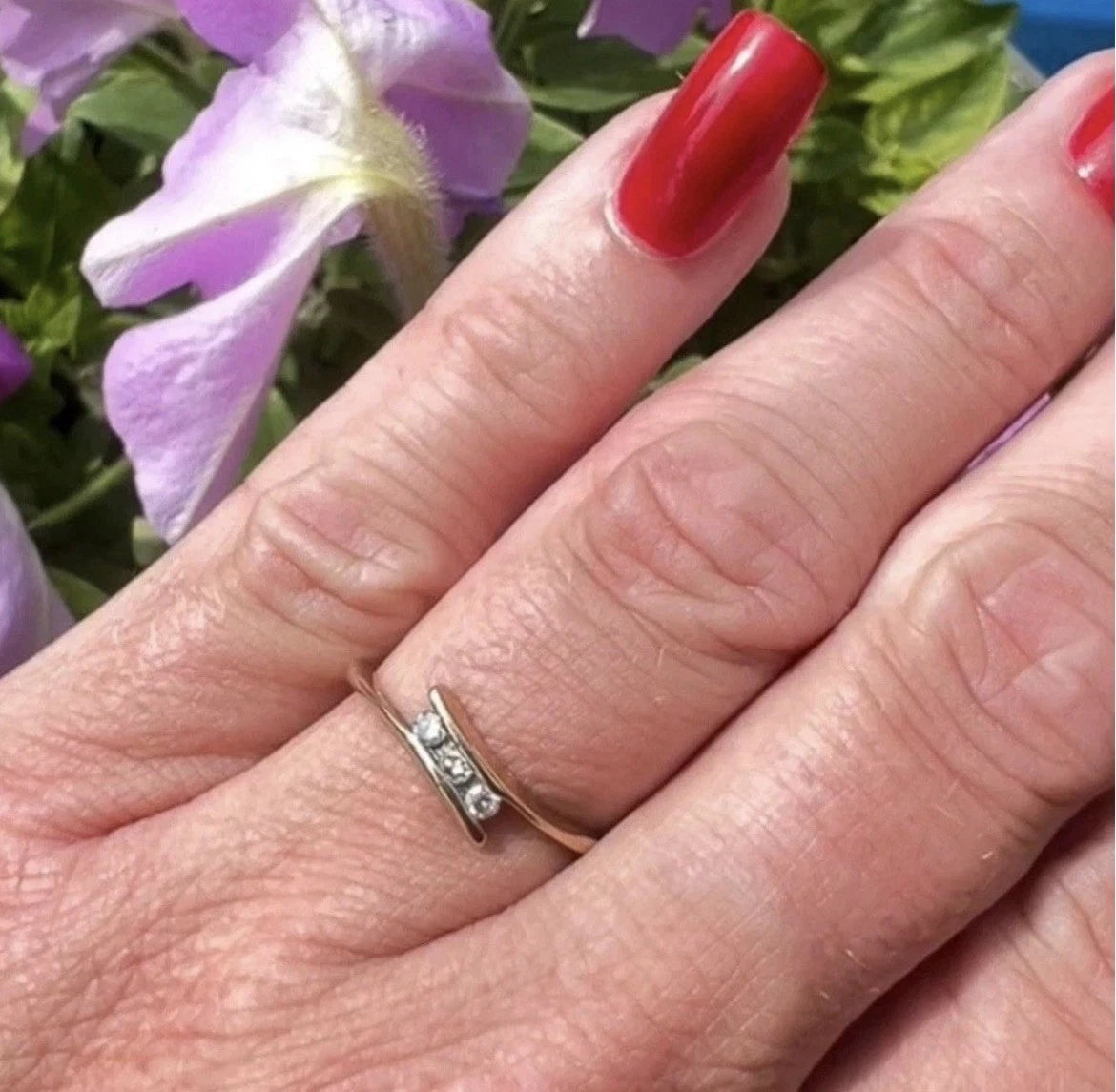 Hand wearing a three stone gold ring with red nail polish against a blurred floral background