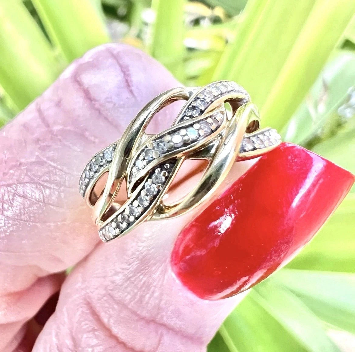 Gold ring with diamonds on a hand with red nail polish against a green leafy background