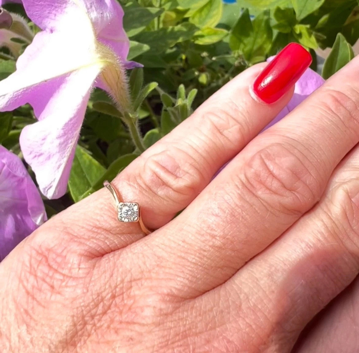Hand with red nail polish wearing a gold ring with a diamond, surrounded by greenery and flowers.