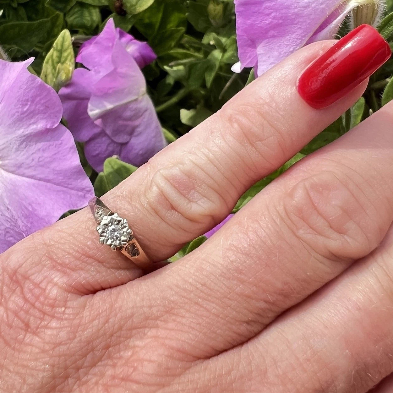 Hand wearing a diamond ring with purple flowers in the background