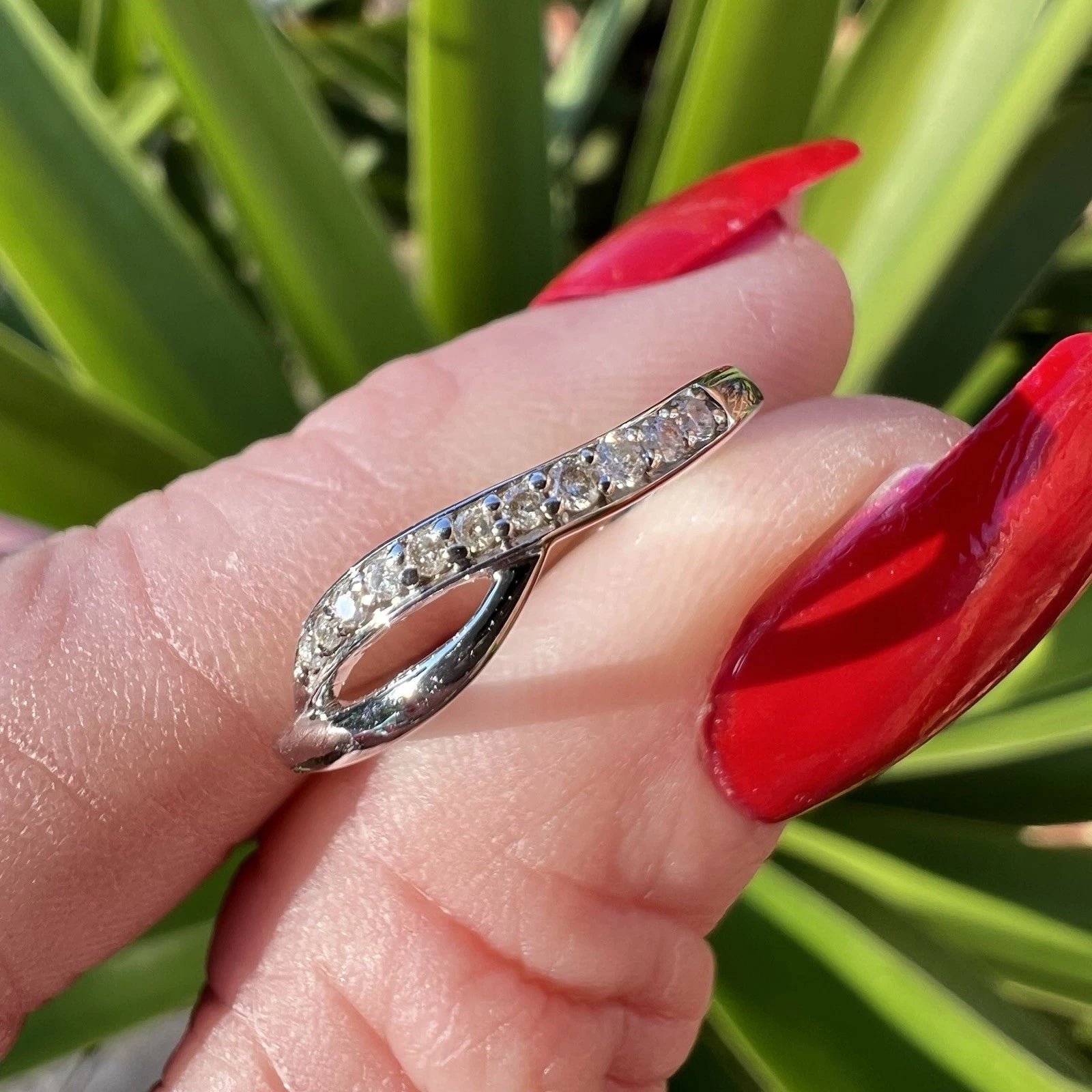 White gold ring with diamonds held by fingers against a red nail varnish  and green leaves background