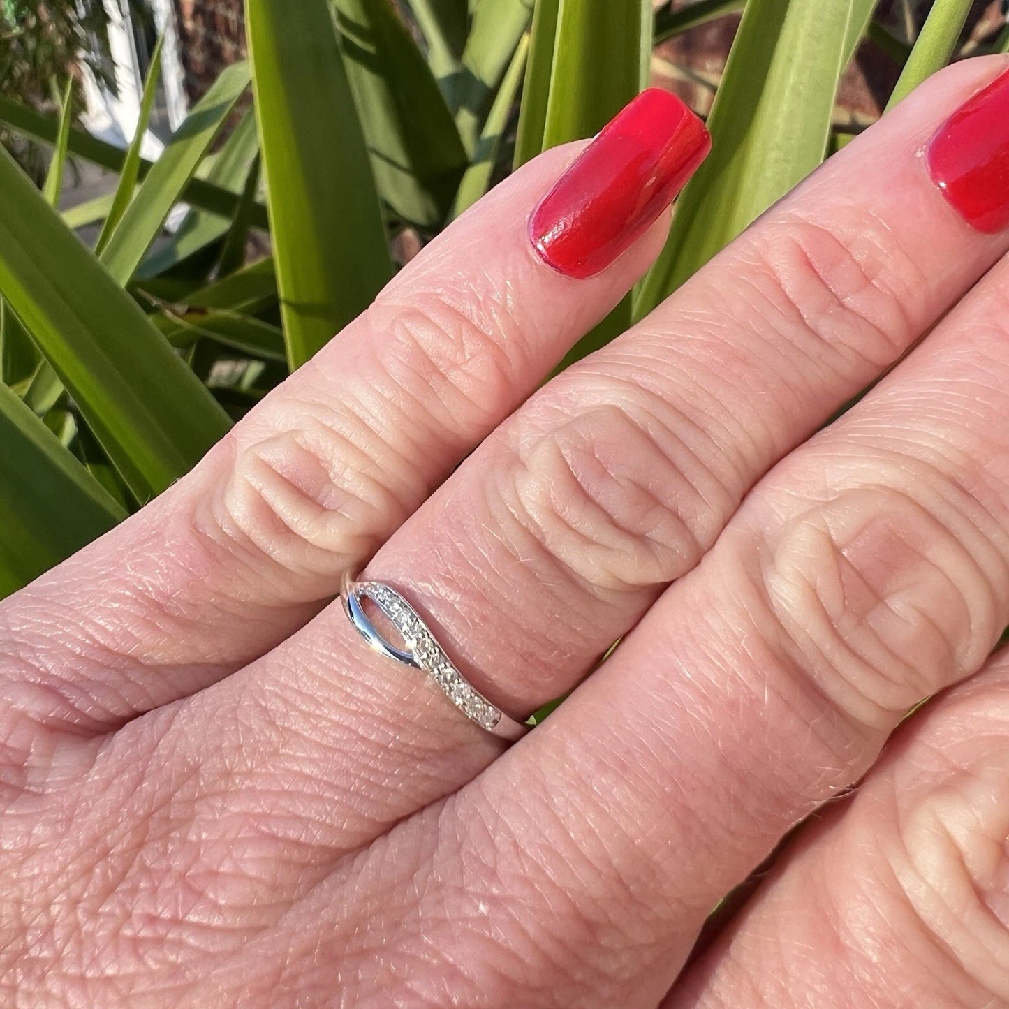 Hand wearing a white gold ring with a diamond, red nail polish against green leaves.