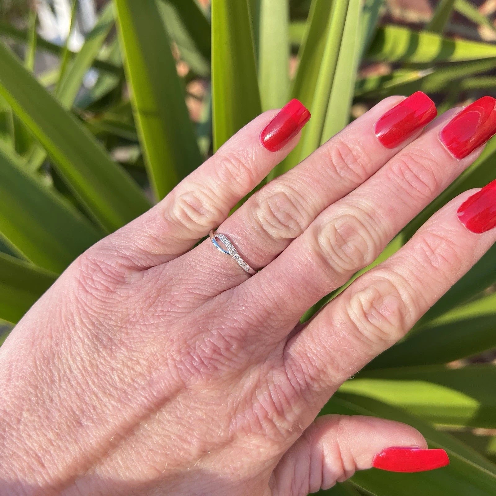 Hand with red nail polish wearing a white gold ring against a green leafy background
