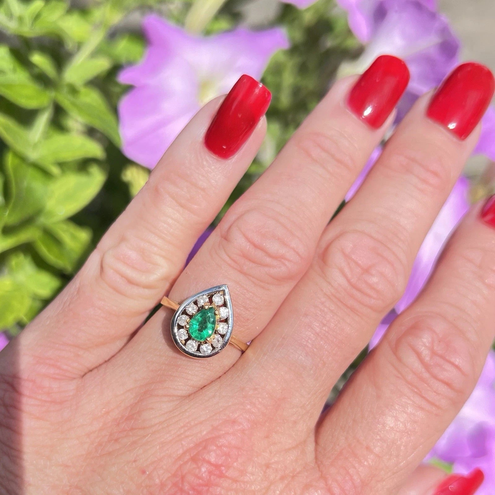 Hand wearing a ring with a green emerald gemstone and diamond halo against a blurred floral background