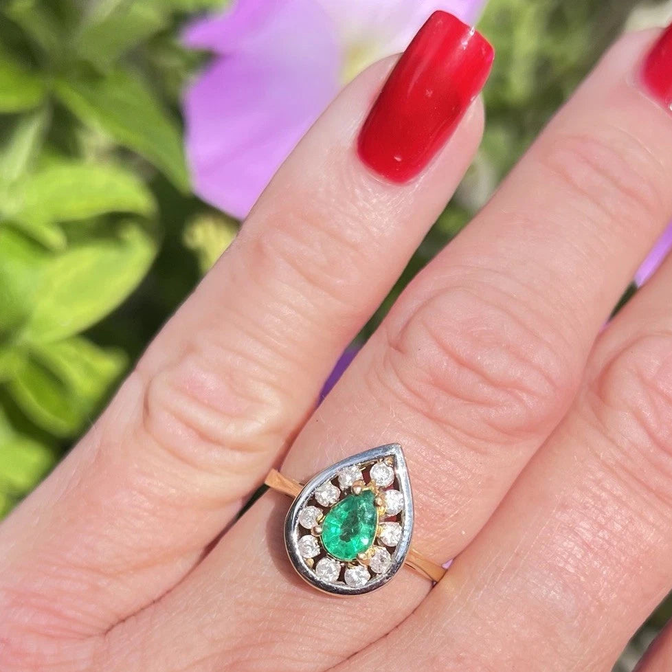 Ring with a green emerald gemstone and diamond halo on a finger with red nail polish against a blurred floral background