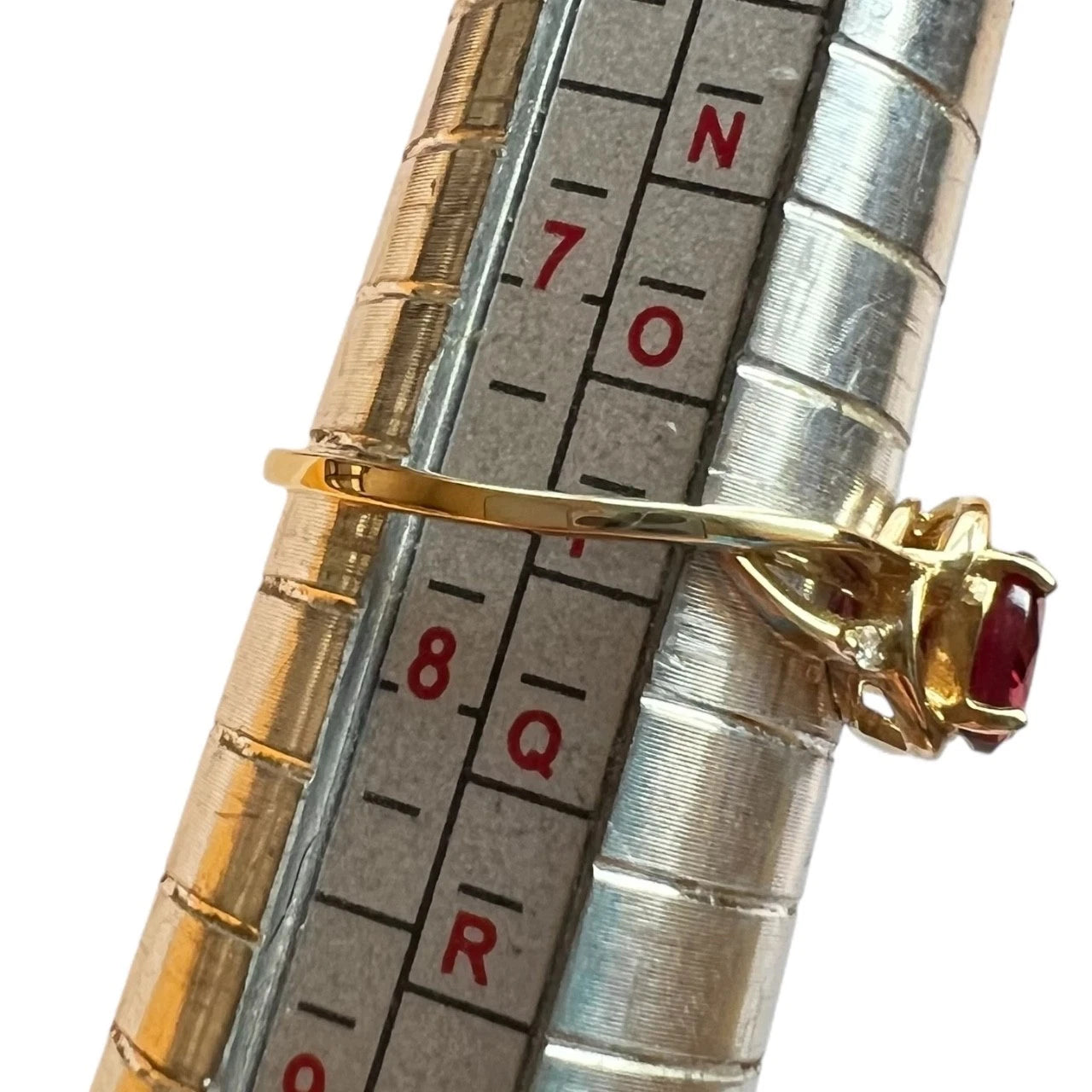 Close-up of a metallic ring sizer with a gold ring and red gemstone on a white background