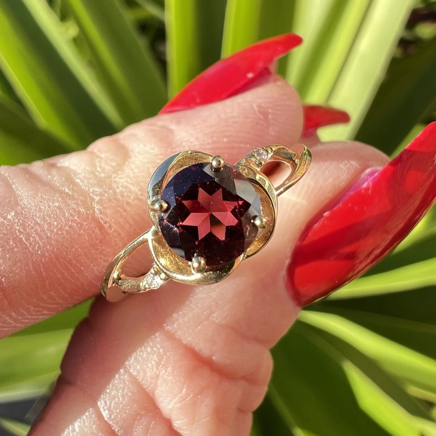 Gold ring with a colourful red garnet gemstone and diamond assents held by a hand against a green and red leafy background