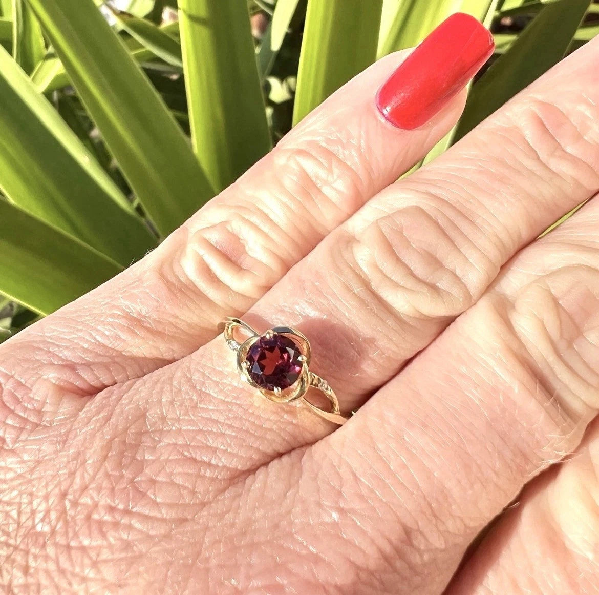 Hand wearing a gold ring with a red gemstone against a green leafy background