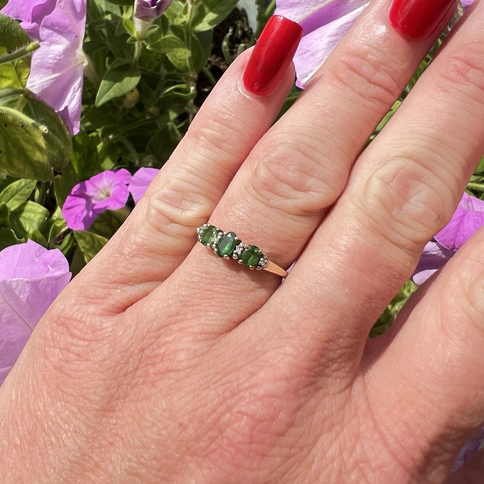 Hand wearing a ring with green gemstones against a background of purple flowers