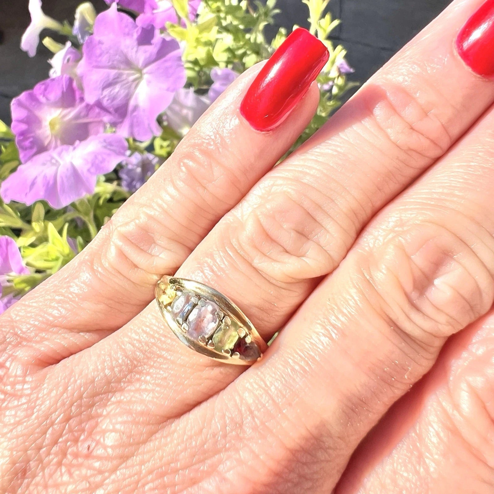Hand wearing a gold ring with a multicolored gemstone, red nails, and purple flowers in the background.